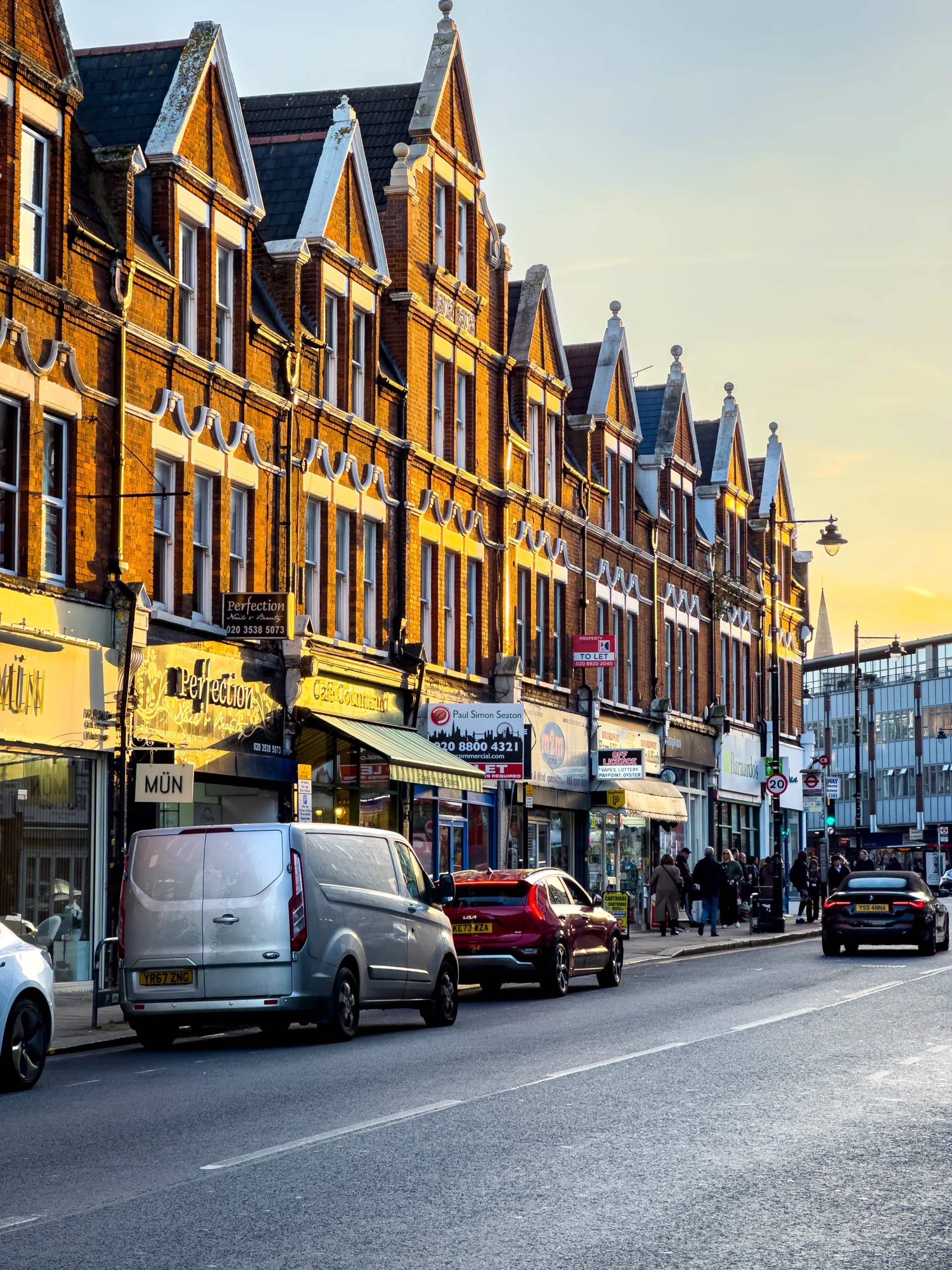 Busy high street lined with red-brick Victorian-style buildings, shops, and parked cars