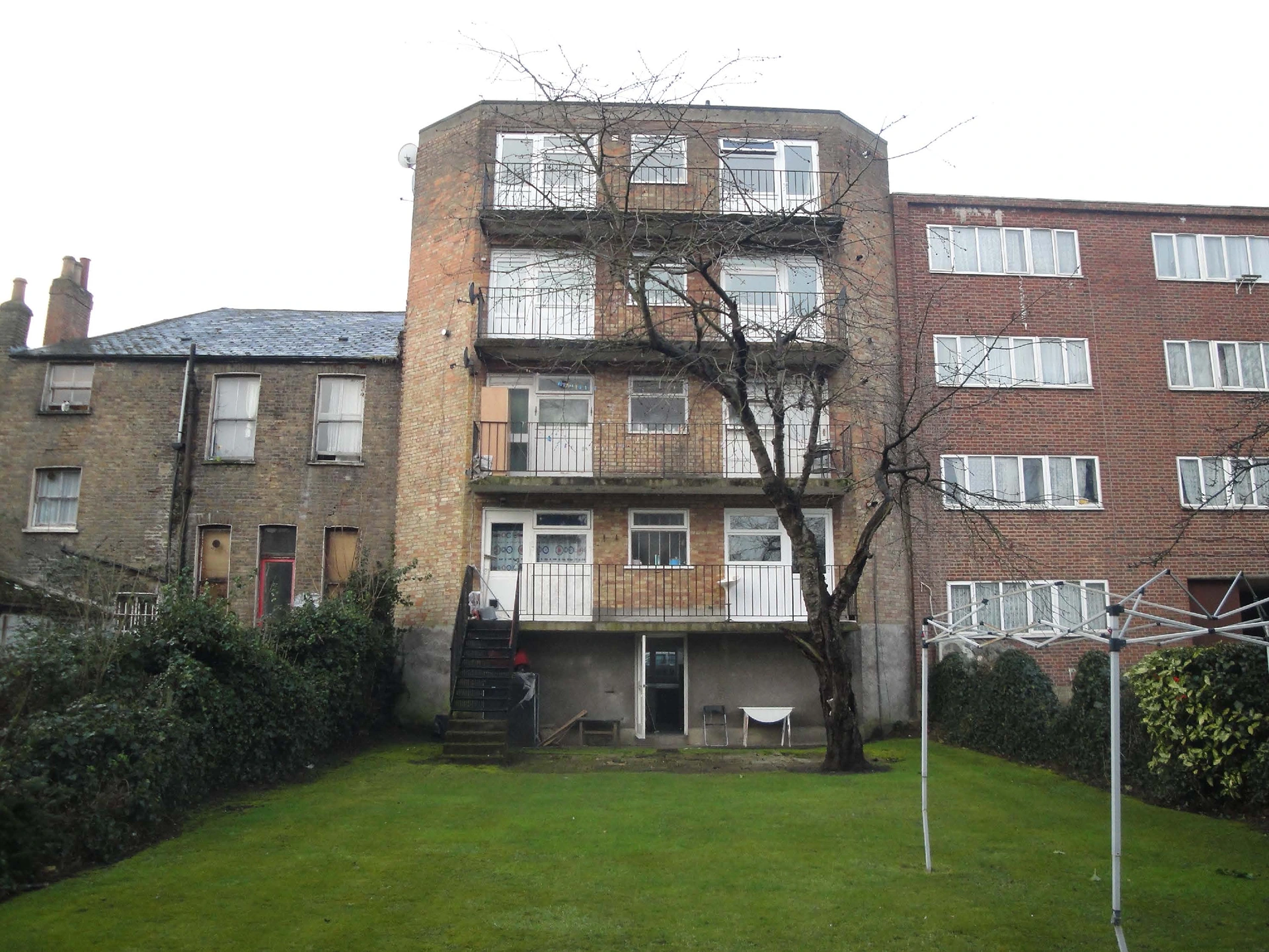 Rear view of an old brick apartment building with worn balconies and an overgrown garden before insulation upgrade