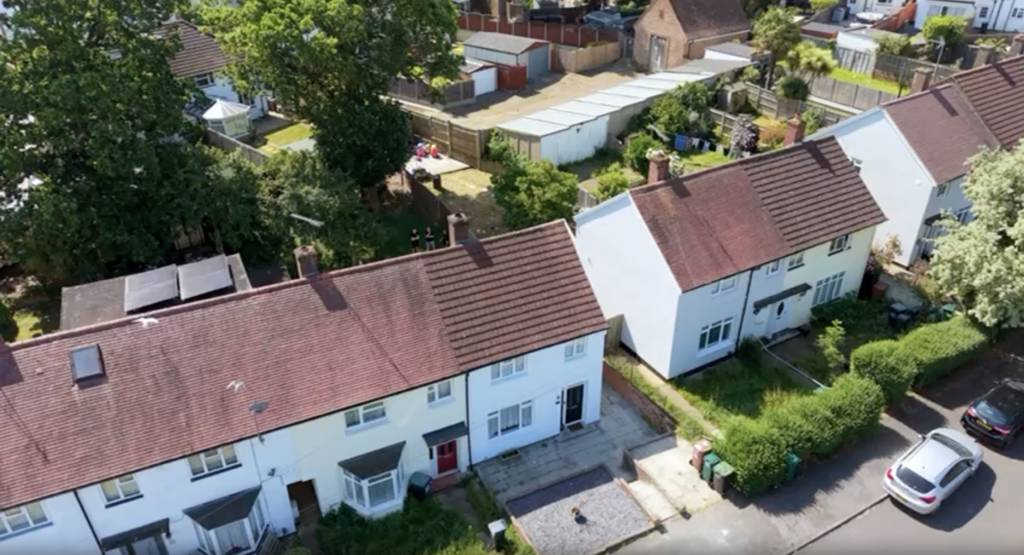 An aerial, angled photograph displays a row of two-story white terraced houses with red-tiled roofs, surrounded by green trees, backyard gardens, and a car parked on the street.
