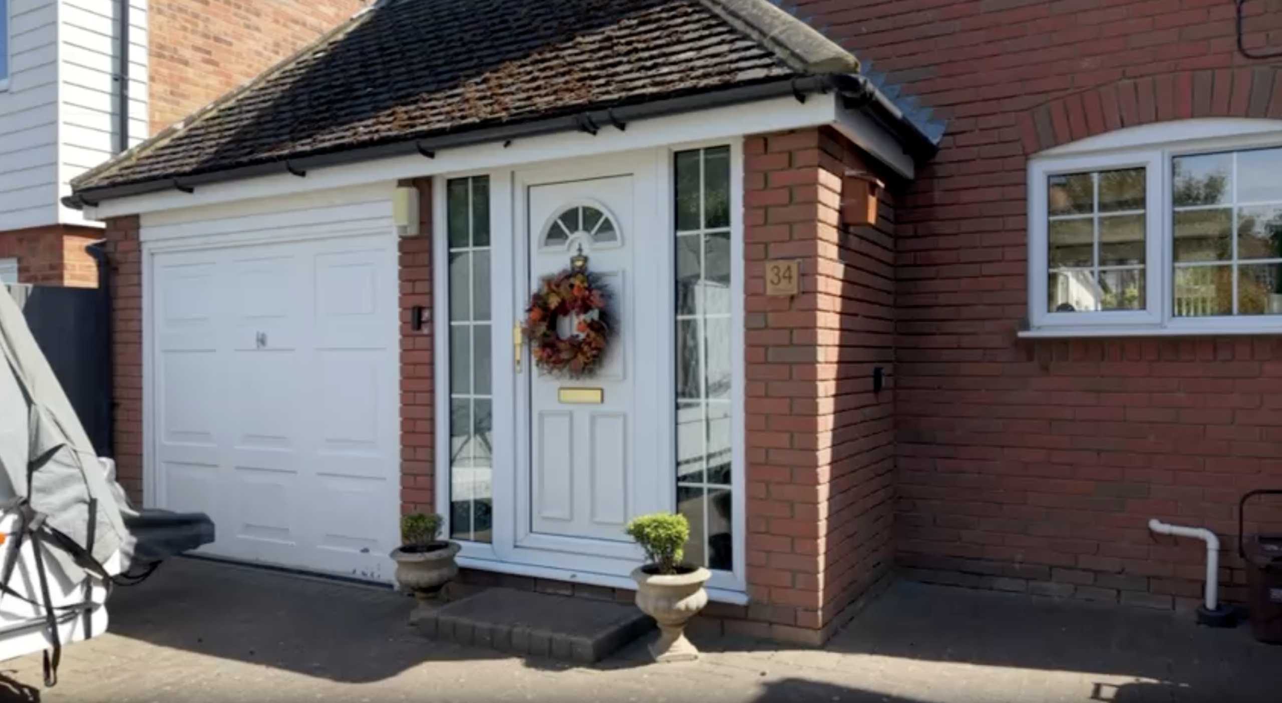 An exterior shot of a modern red brick house in Baldock, featuring a white front door with sidelights, a white garage door to its left, and a multi-panel window to its right. A decorative autumn wreath hangs on the front door, flanked by two potted plants on the pavement.