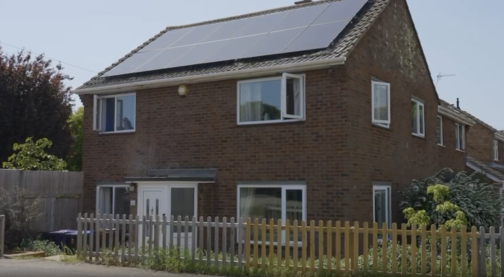 A two-story brick house with a fenced front yard and solar panels installed across the entire roof.
