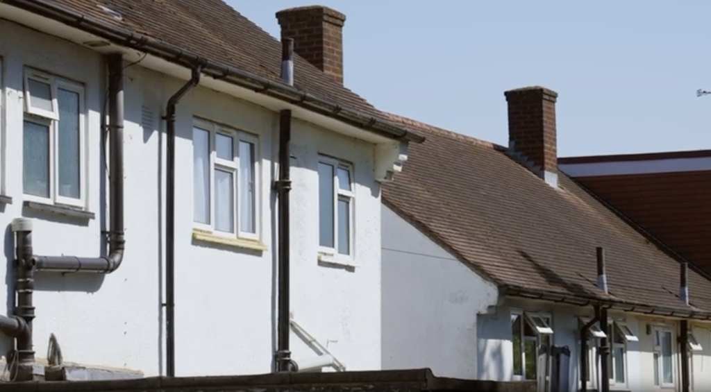 A white building with windows, a brown roof, two brick chimneys, and black drainpipes.