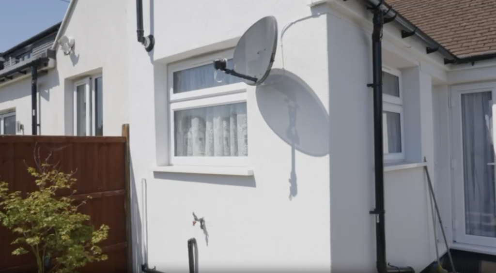 A close-up of a white building with windows and a satellite dish mounted on the exterior wall next to a wooden fence.