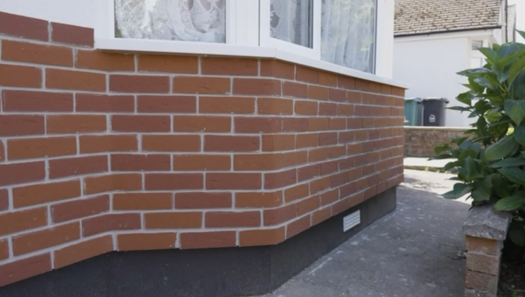 A house exterior featuring a freshly installed brick-slip wall beneath a white bay window, with a dark foundation base and a paved walkway alongside green bushes.