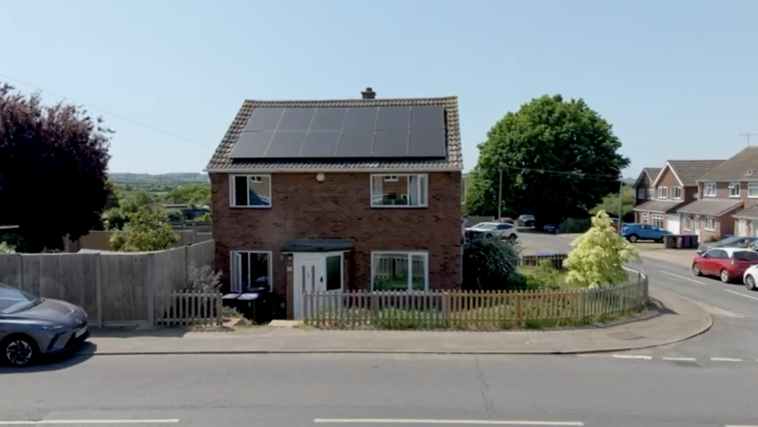 An eye-level, wide shot shows a two-story red brick house in Hertfordshire with a large solar panel array covering its roof under a clear, bright blue sky.