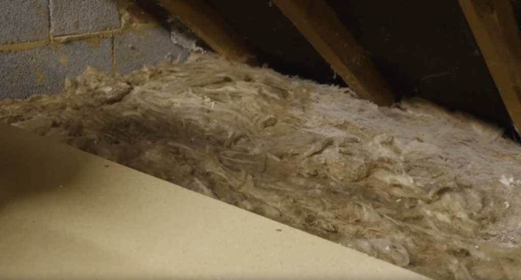 An image showing a close-up, angled view inside an attic space. Fluffy, light-brown mineral wool or fiberglass insulation fills the floor cavity between wooden roof rafters and a flat floorboard surface.